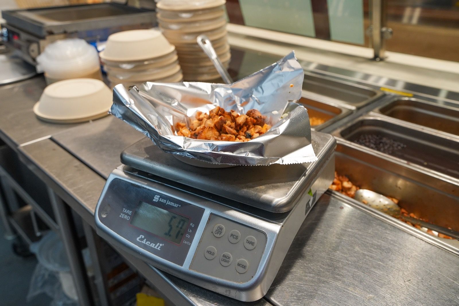 A foil-lined bowl of chicken on a digital kitchen scale, beside the steel pans of a food line